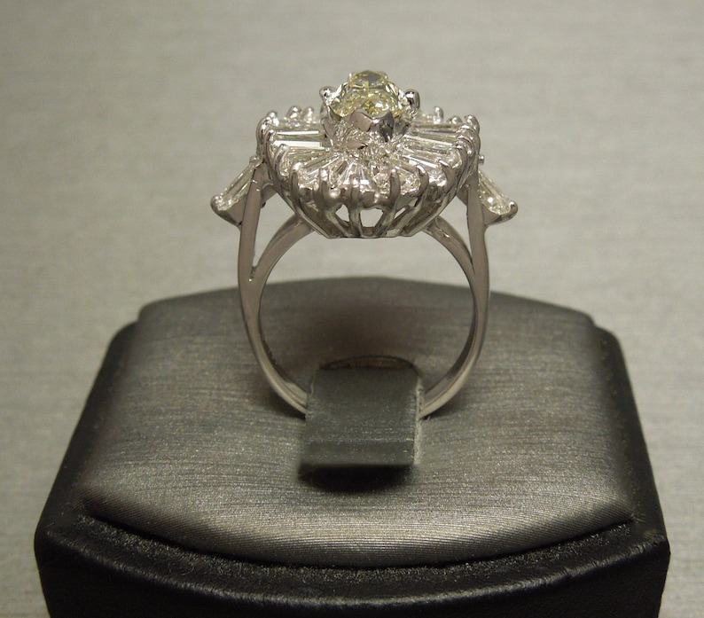 Op de afbeelding: Een zilveren ring met een grote, ovale, gele edelsteen omringd door kleinere, baguettevormige diamanten.