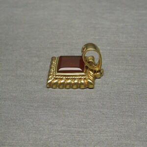 May include: A gold pendant with a square red carnelian gemstone set in a decorative frame. The pendant has a loop for attaching to a chain.