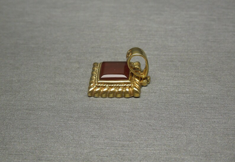 May include: A gold pendant with a square red carnelian gemstone set in a decorative frame. The pendant has a loop for attaching to a chain.