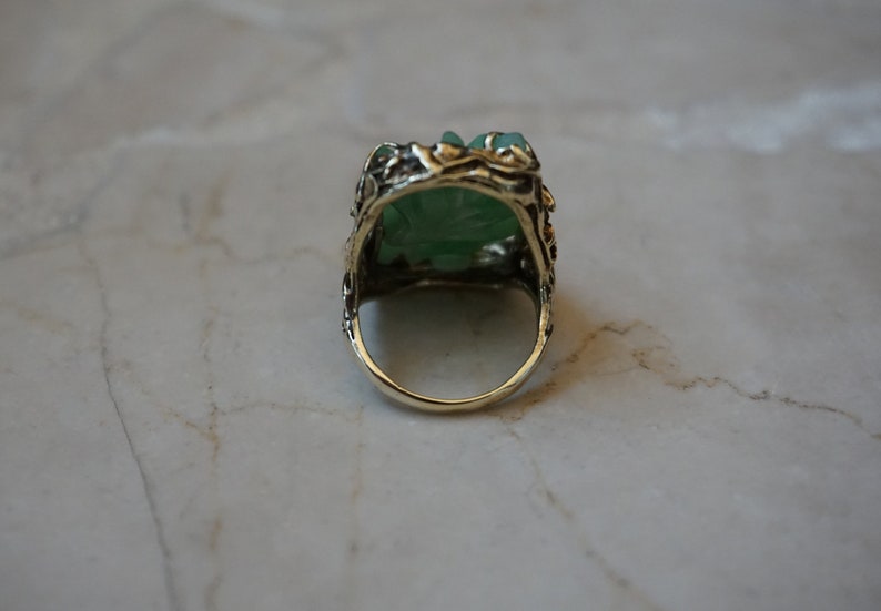 May include: A green jade ring with a detailed gold setting. The ring is set with a carved jade stone and has a delicate floral design.