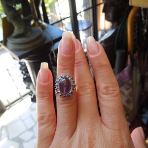May include: Oval amethyst ring with a halo of small blue gemstones. The ring is set on a silver band and is displayed on a hand with long, manicured nails. The ring's central stone is a deep purple color.