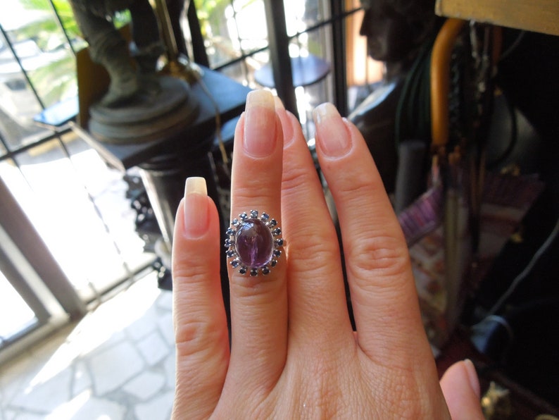 May include: Oval amethyst ring with a halo of small blue gemstones. The ring is set on a silver band and is displayed on a hand with long, manicured nails. The ring's central stone is a deep purple color.