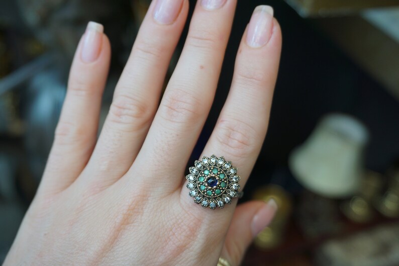 Pu&ograve; includere: Un anello d'argento con una pietra centrale rotonda circondata da pietre pi&ugrave; piccole. La pietra centrale &egrave; uno zaffiro blu scuro, circondato da un anello di pietre verdi e poi da un anello di pietre bianche.