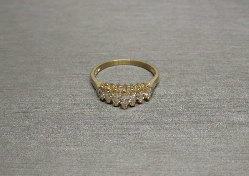 May include: A gold ring featuring a series of marquise-cut gemstones. The gemstones are set in a row across the top of the ring. The ring is set against a neutral gray background.