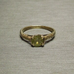 May include: A gold ring with a green gemstone set in a prong setting. The band is decorated with small, raised lines.