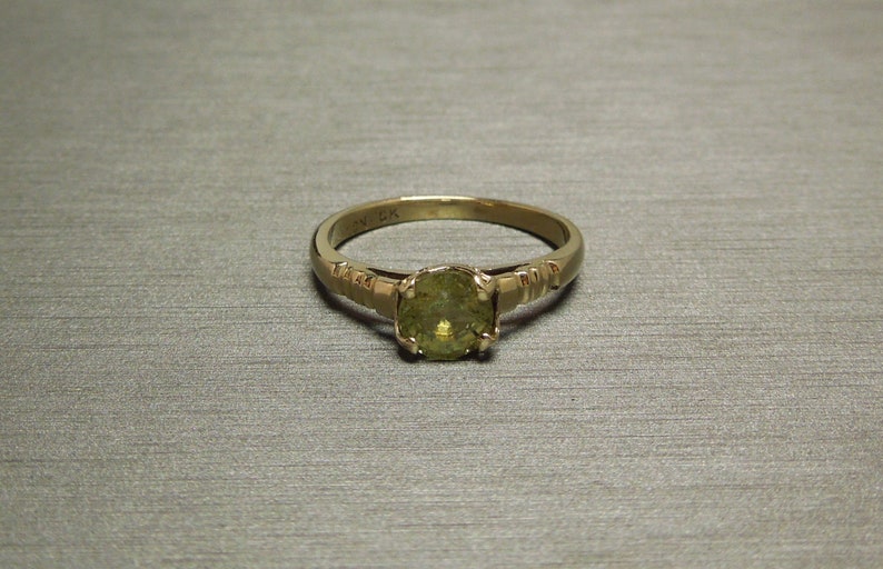 May include: A gold ring with a green gemstone set in a prong setting. The band is decorated with small, raised lines.