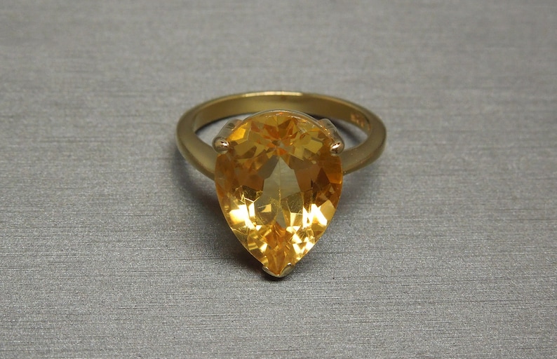 May include: A gold ring featuring a large pear-shaped topaz gemstone. The gemstone is a bright yellow colour.