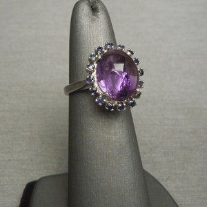 May include: A silver ring featuring a large, oval-shaped purple gemstone, surrounded by a halo of smaller blue gemstones. The ring is displayed on a gray stand, showcasing its design and color contrast.