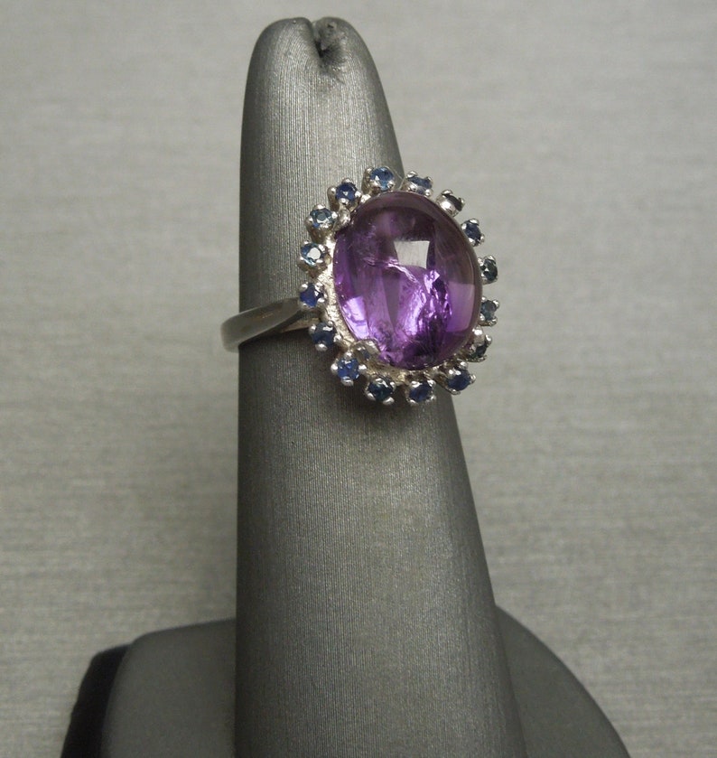 May include: A silver ring featuring a large, oval-shaped purple gemstone, surrounded by a halo of smaller blue gemstones. The ring is displayed on a gray stand, showcasing its design and color contrast.