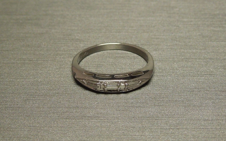 May include: A silver band ring with a row of small, rectangular-shaped diamonds set in the center.