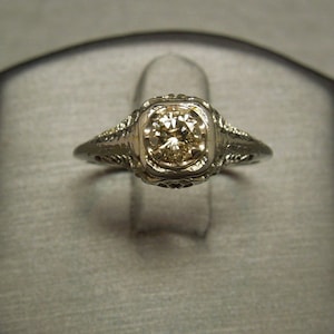Puede incluir: Un anillo de compromiso vintage con un delicado dise&ntilde;o de filigrana y un diamante redondo y transparente.