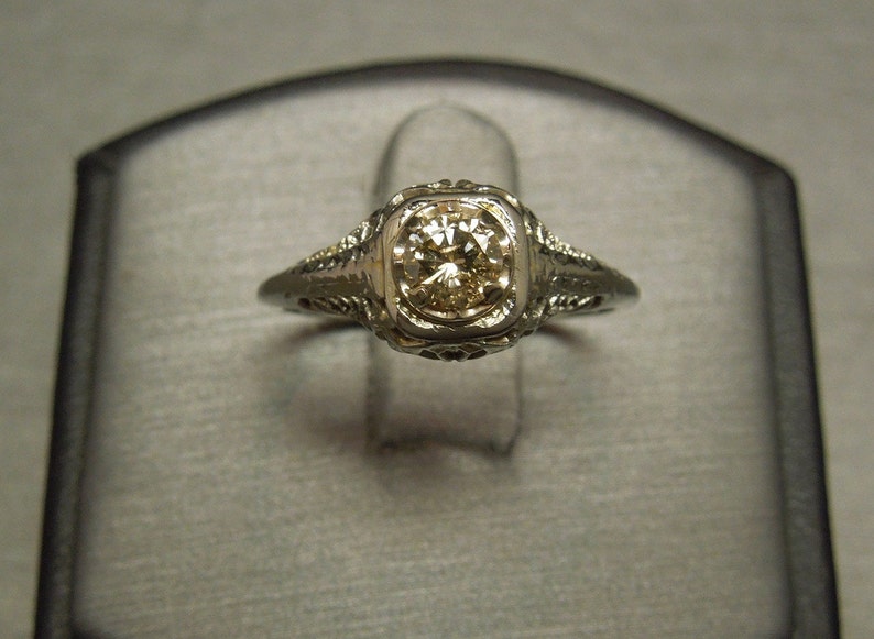Puede incluir: Un anillo de compromiso vintage con un delicado dise&ntilde;o de filigrana y un diamante redondo y transparente.