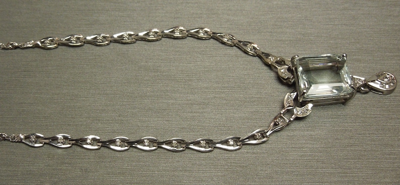 K&ouml;nnte beinhalten: Eine silberne Halskette mit einem gro&szlig;en, rechteckigen, hellblauen Edelsteinanh&auml;nger. Der Anh&auml;nger ist von kleineren Diamanten umgeben. Die Kette hat ein filigranes Design mit kleinen, tropfenf&ouml;rmigen Gliedern, jeweils mit einem kleinen Diamanten.
