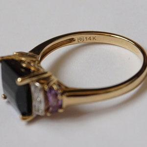 Pu&ograve; includere: Un anello d'oro con una grande pietra quadrata nera, due pietre viola pi&ugrave; piccole e una piccola fila di pietre trasparenti. L'anello &egrave; contrassegnato "14K" sulla fascia interna.