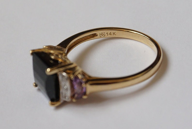 Pu&ograve; includere: Un anello d'oro con una grande pietra quadrata nera, due pietre viola pi&ugrave; piccole e una piccola fila di pietre trasparenti. L'anello &egrave; contrassegnato "14K" sulla fascia interna.