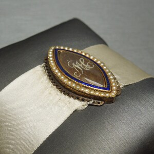 May include: A gold locket with a blue enamel border and a pearl trim. The locket has a clear glass insert with the initials "M.C." written in script inside.