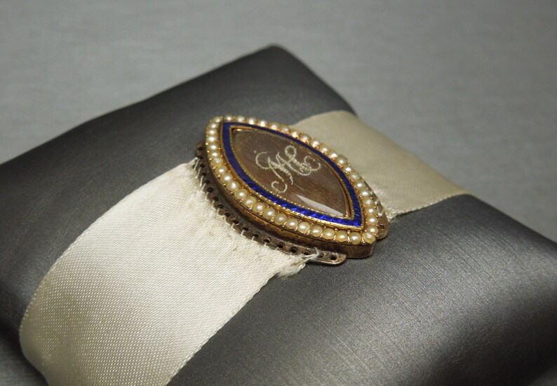 May include: A gold locket with a blue enamel border and a pearl trim. The locket has a clear glass insert with the initials "M.C." written in script inside.