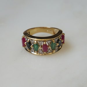 May include: A gold ring with a band of small diamonds and five oval-shaped gemstones. The gemstones are red, green, and blue.