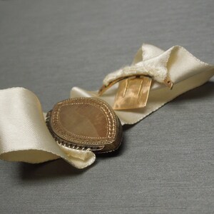 May include: A gold locket with a decorative border and a white satin ribbon. The locket is closed and has a clasp on the back.