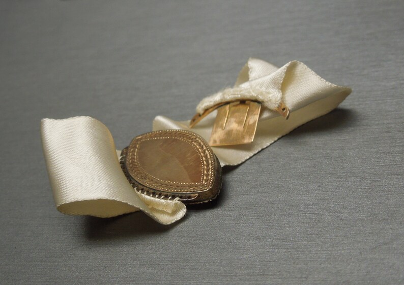 May include: A gold locket with a decorative border and a white satin ribbon. The locket is closed and has a clasp on the back.
