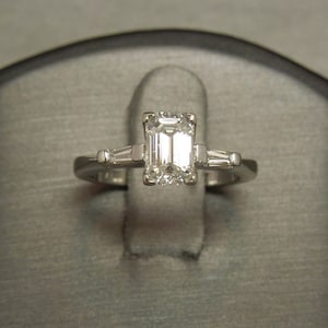 May include: A silver ring with an emerald-cut diamond center stone and two smaller baguette-cut diamonds on either side.