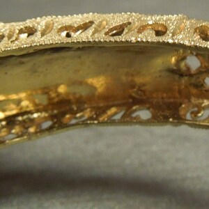 May include: Gold-tone metal bracelet with an intricate filigree design. The bracelet has a hinged clasp and is decorated with small, raised details.