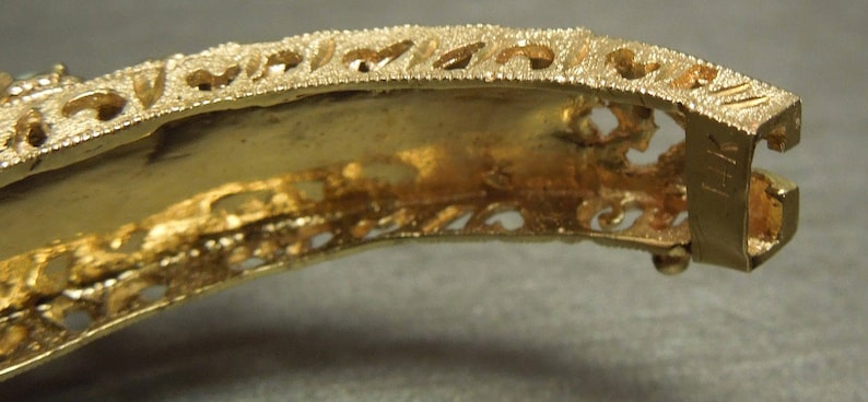 May include: Gold-tone metal bracelet with an intricate filigree design. The bracelet has a hinged clasp and is decorated with small, raised details.