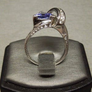 May include: A silver ring with a large, pear-shaped blue gemstone and a band of smaller diamonds. The ring is on a gray velvet ring box.