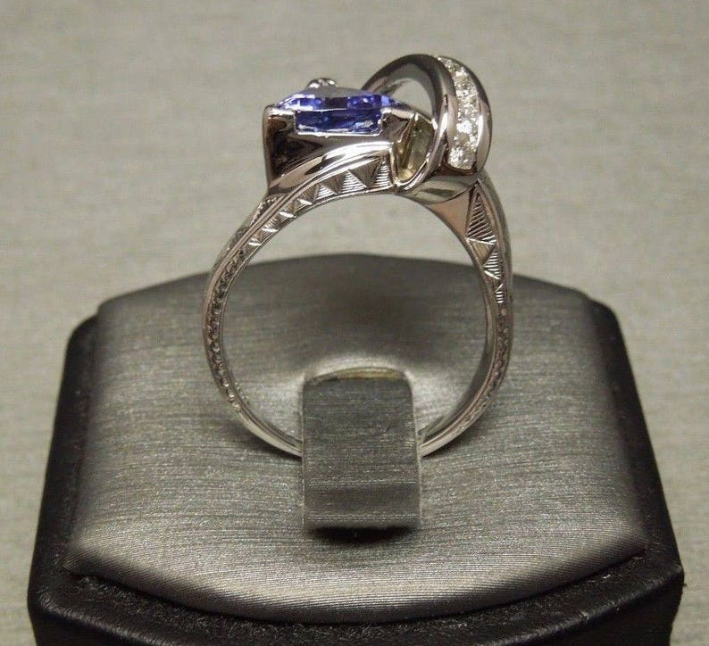 May include: A silver ring with a large, pear-shaped blue gemstone and a band of smaller diamonds. The ring is on a gray velvet ring box.