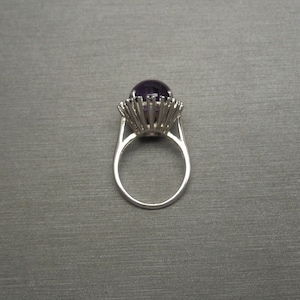 May include: A silver ring featuring a large, round, deep purple gemstone. The gemstone is set in a detailed, spiky setting, and the ring has a simple, elegant band. The ring is shown against a gray background.