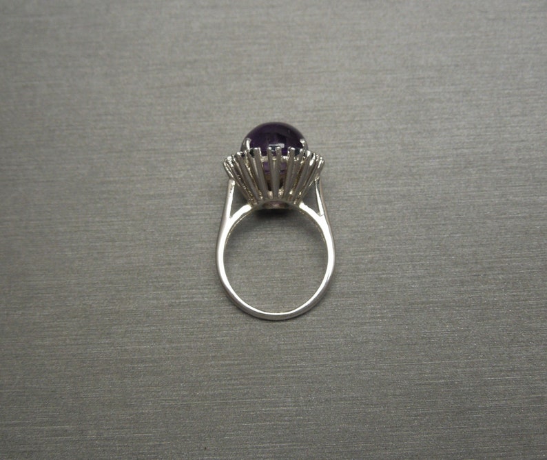 May include: A silver ring featuring a large, round, deep purple gemstone. The gemstone is set in a detailed, spiky setting, and the ring has a simple, elegant band. The ring is shown against a gray background.