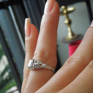 May include: A silver ring with a large, clear, rectangular-shaped diamond set in a delicate, ornate design.