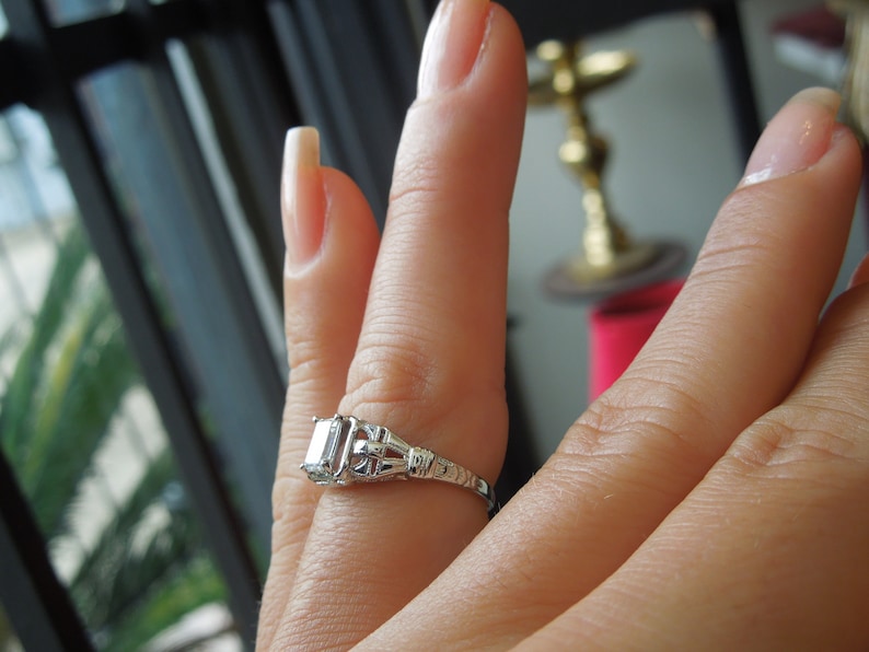 May include: A silver ring with a large, clear, rectangular-shaped diamond set in a delicate, ornate design.