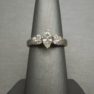 Pode incluir: Uma alian&ccedil;a de noivado em ouro branco com um diamante central em forma de marquise e dois diamantes redondos menores de cada lado. A banda &eacute; gravada com um delicado desenho floral.
