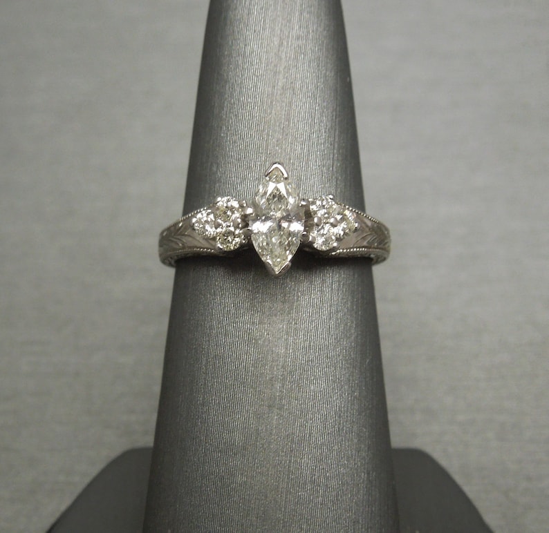 Pode incluir: Uma alian&ccedil;a de noivado em ouro branco com um diamante central em forma de marquise e dois diamantes redondos menores de cada lado. A banda &eacute; gravada com um delicado desenho floral.