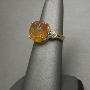 May include: A gold ring with a carved amber stone and a small diamond accent. The ring is on a gray display stand.