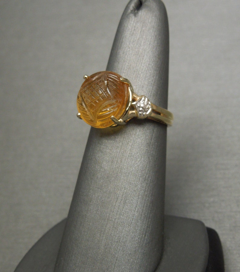 May include: A gold ring with a carved amber stone and a small diamond accent. The ring is on a gray display stand.