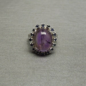 May include: Oval amethyst ring with a halo of small, dark blue gemstones. The ring has a silver band and a large, polished purple amethyst center stone. The gemstones are set around the amethyst, creating a floral-like design.
