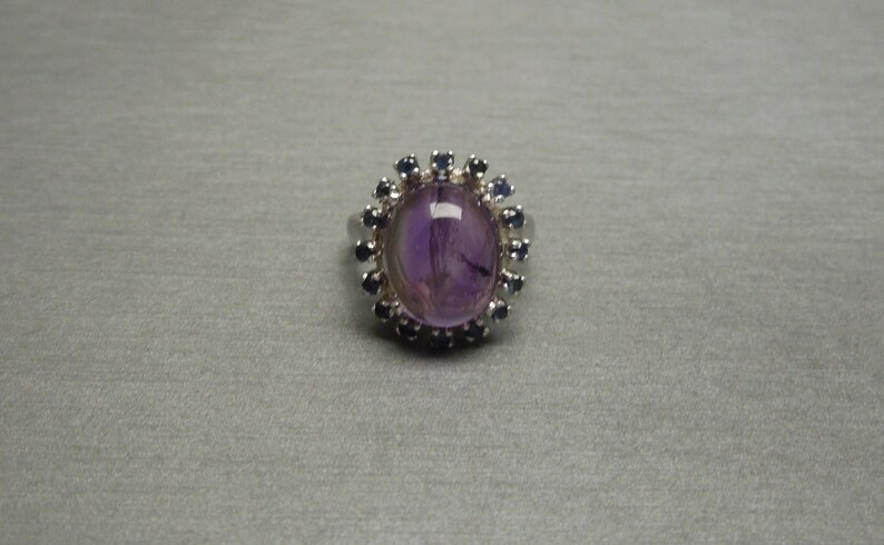 May include: Oval amethyst ring with a halo of small, dark blue gemstones. The ring has a silver band and a large, polished purple amethyst center stone. The gemstones are set around the amethyst, creating a floral-like design.