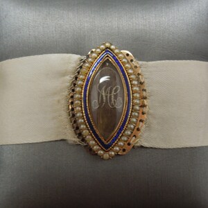 May include: A gold locket with a blue enamel border and a pearl trim. The locket contains a lock of hair with the initials "MC" woven into it.