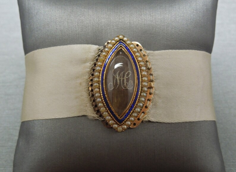 May include: A gold locket with a blue enamel border and a pearl trim. The locket contains a lock of hair with the initials "MC" woven into it.