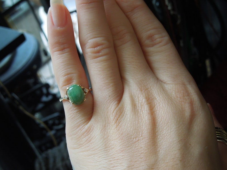 K&ouml;nnte beinhalten: Ein goldener Ring mit einem gr&uuml;nen ovalen Edelstein und kleineren Diamanten auf der Ringschiene.
