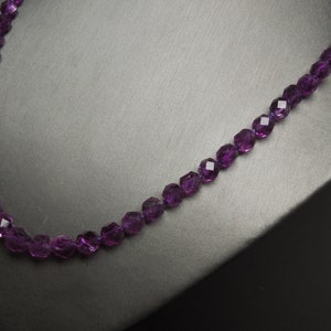 Pode incluir: Um colar de pedras preciosas roxas com contas facetadas. O colar est&aacute; sobre um fundo cinza.