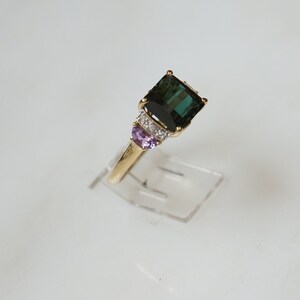 Pu&ograve; includere: Un anello d'oro con una grande pietra preziosa verde smeraldo circondata da pietre preziose pi&ugrave; piccole, chiare e viola.