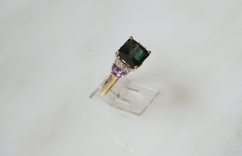 Pu&ograve; includere: Un anello d'oro con una grande pietra preziosa verde smeraldo circondata da pietre preziose pi&ugrave; piccole, chiare e viola.
