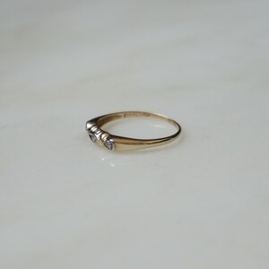 May include: A gold ring featuring three small, clear gemstones. The ring is set on a white surface. The band is thin and polished. The gemstones are round and closely set together.