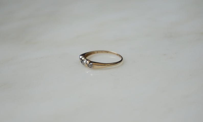 May include: A gold ring featuring three small, clear gemstones. The ring is set on a white surface. The band is thin and polished. The gemstones are round and closely set together.