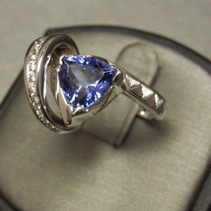 May include: A silver ring with a large, triangular, purple gemstone surrounded by smaller diamonds. The ring is set on a black velvet background.