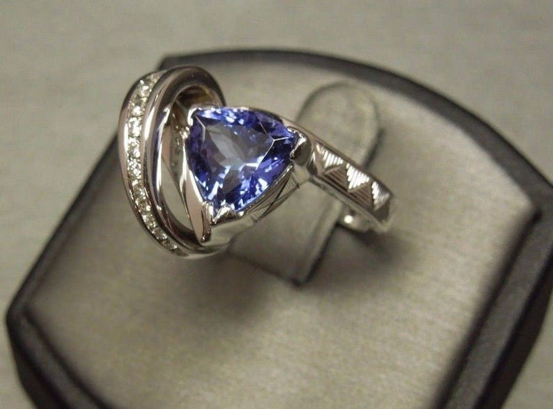 May include: A silver ring with a large, triangular, purple gemstone surrounded by smaller diamonds. The ring is set on a black velvet background.