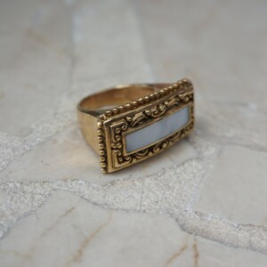 May include: A gold ring with a rectangular white stone set in a detailed ornate frame.